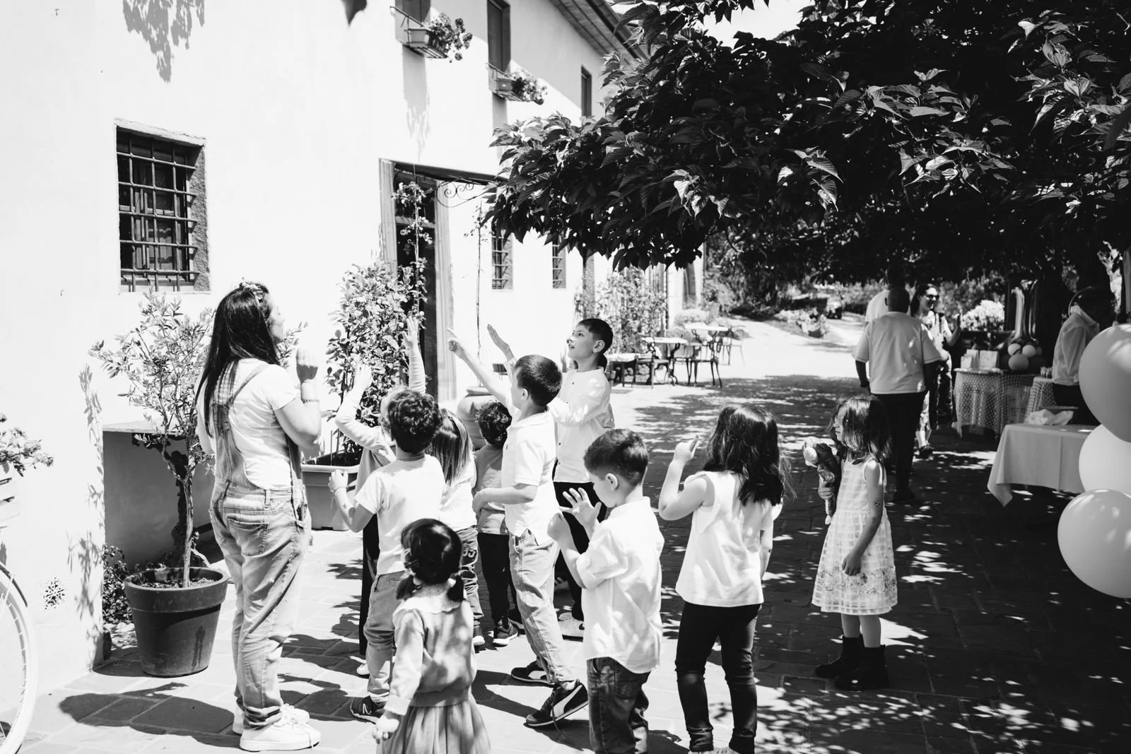 Un gruppo di bambini che giocano e si divertono all'aperto durante una festa in un cortile soleggiato, con alcuni adulti che osservano in sottofondo.