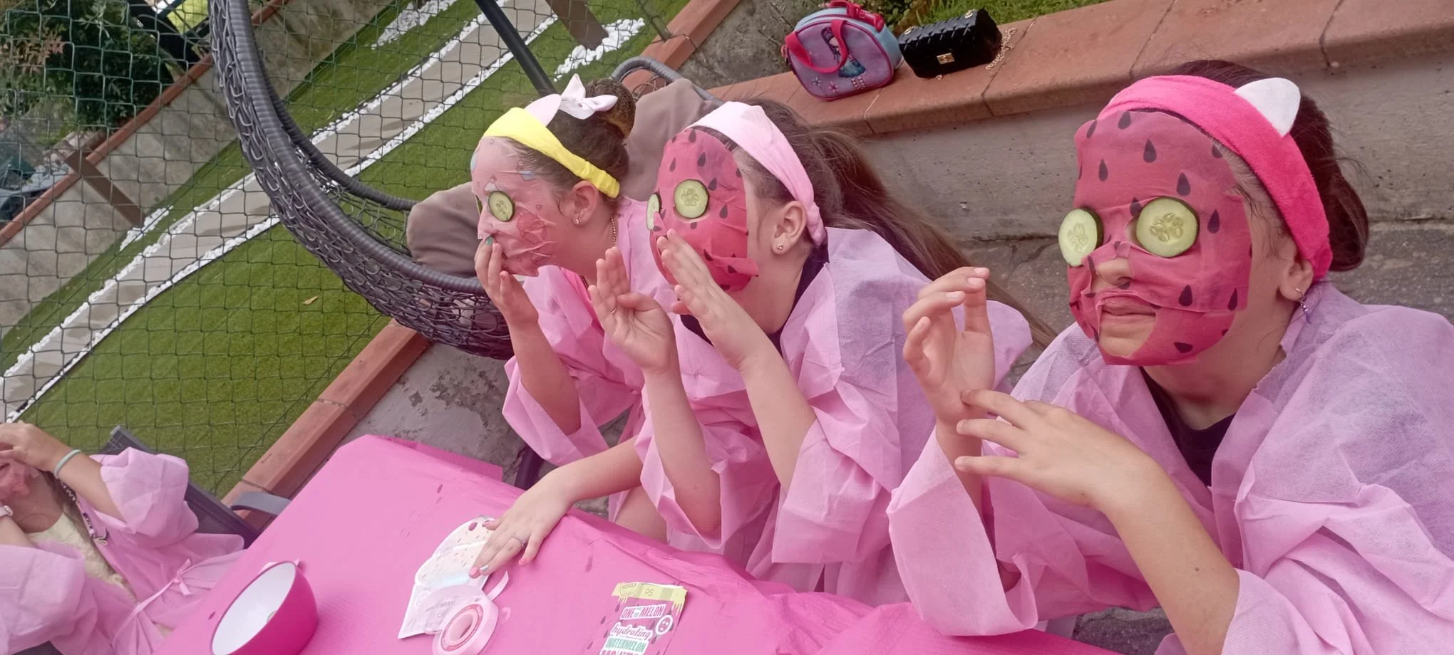 Tre ragazze indossano maschere di viso a forma di anguria e tappi di cetriolo sugli occhi, sedute a un tavolo rosa all'aperto, che sembra fare una festa o un momento di relax.