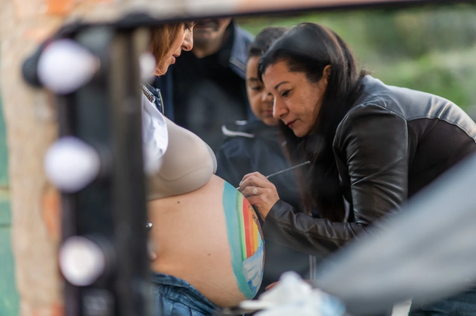 Una donna sta dipingendo un arcobaleno colorato sulla pancia di un'altra donna che è incinta, mentre altre persone osservano.