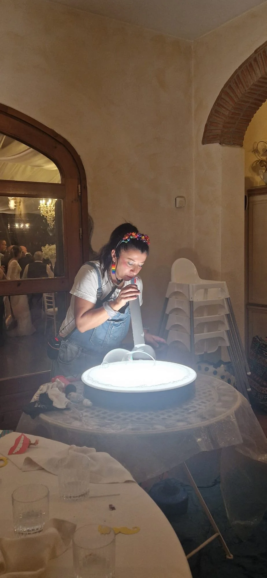 Donna con abbigliamento colorato e acconciatura decorata con fiori, mentre si soffia sopra una torta di compleanno bianca e luminosa su un tavolo coperto con una tovaglia di tulle, in un ristorante con persone sedute al fondo.