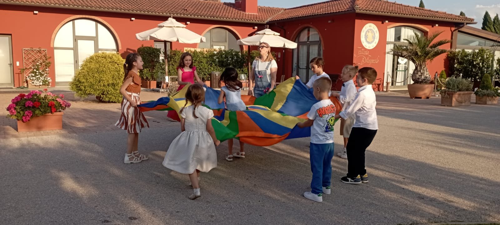 Bambini che giocano con un grande drappo colorato in un cortile esterno con un edificio rosso sullo sfondo, sotto ombrelloni e con piante ornamentali.
