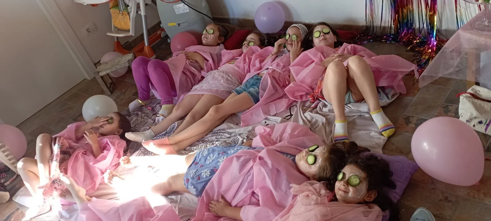 Gruppo di bambine che si rilassano con occhiali da spa, maschere e cuki di cetrioli sul viso, sdraiate su un pavimento decorato con palloncini e festoni, in una festa di compleanno.