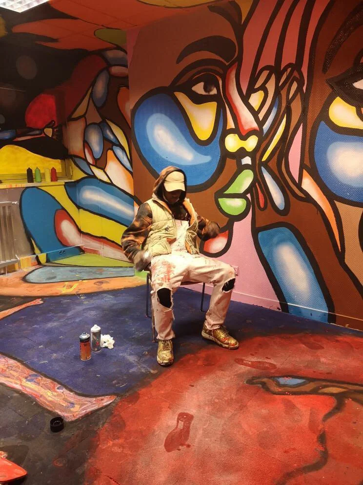 Un artiste de graffiti portant une cagoule, un doudoune et un pantalon déchiré, assis dans une salle ornée de fresques murales colorées et abstraites.