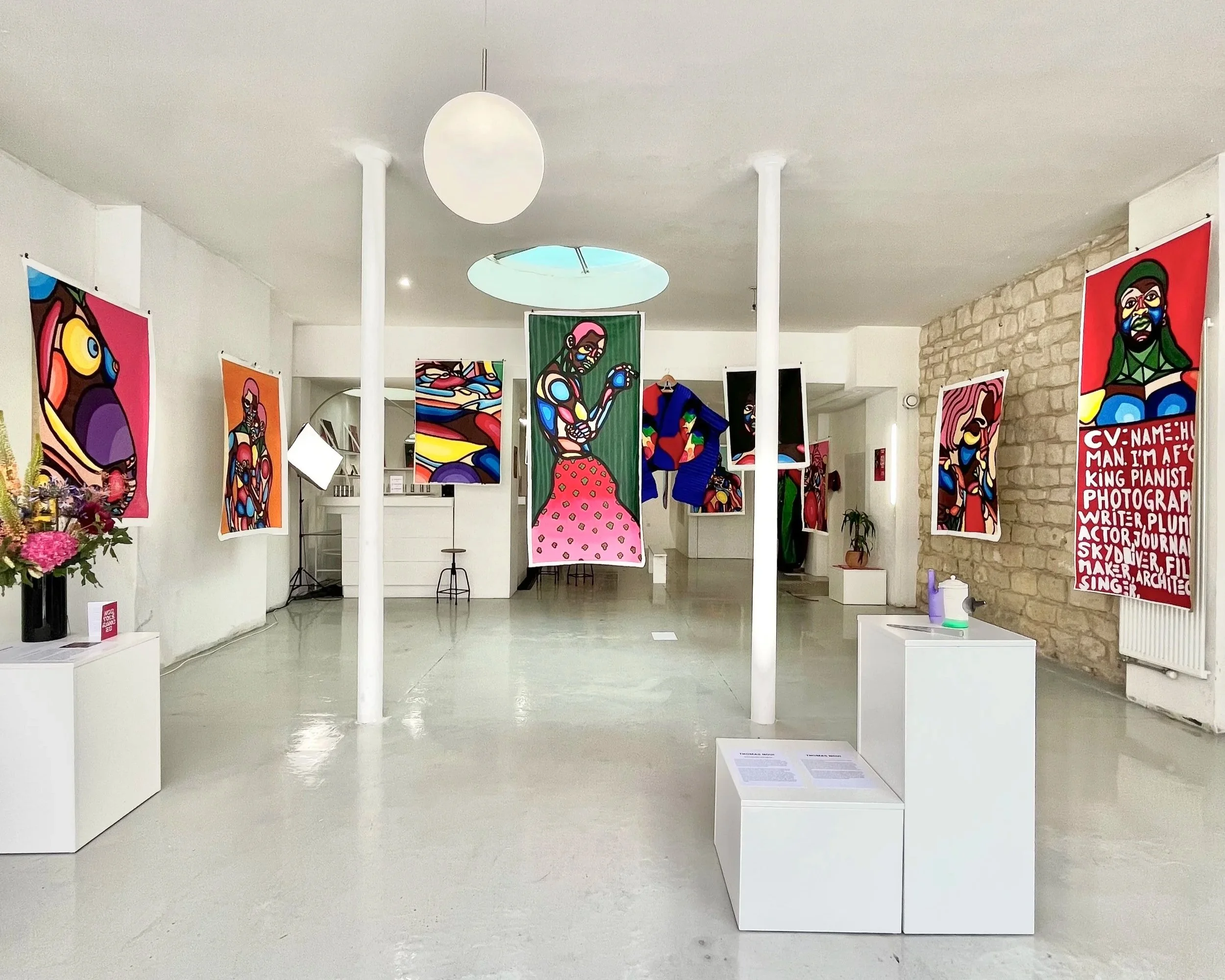 Galerie d'art présentant des œuvres d'art colorées en style abstrait et figuratif, avec plusieurs toiles suspendues sur murs blanc et pierre, et une table avec des fleurs en arrangement.