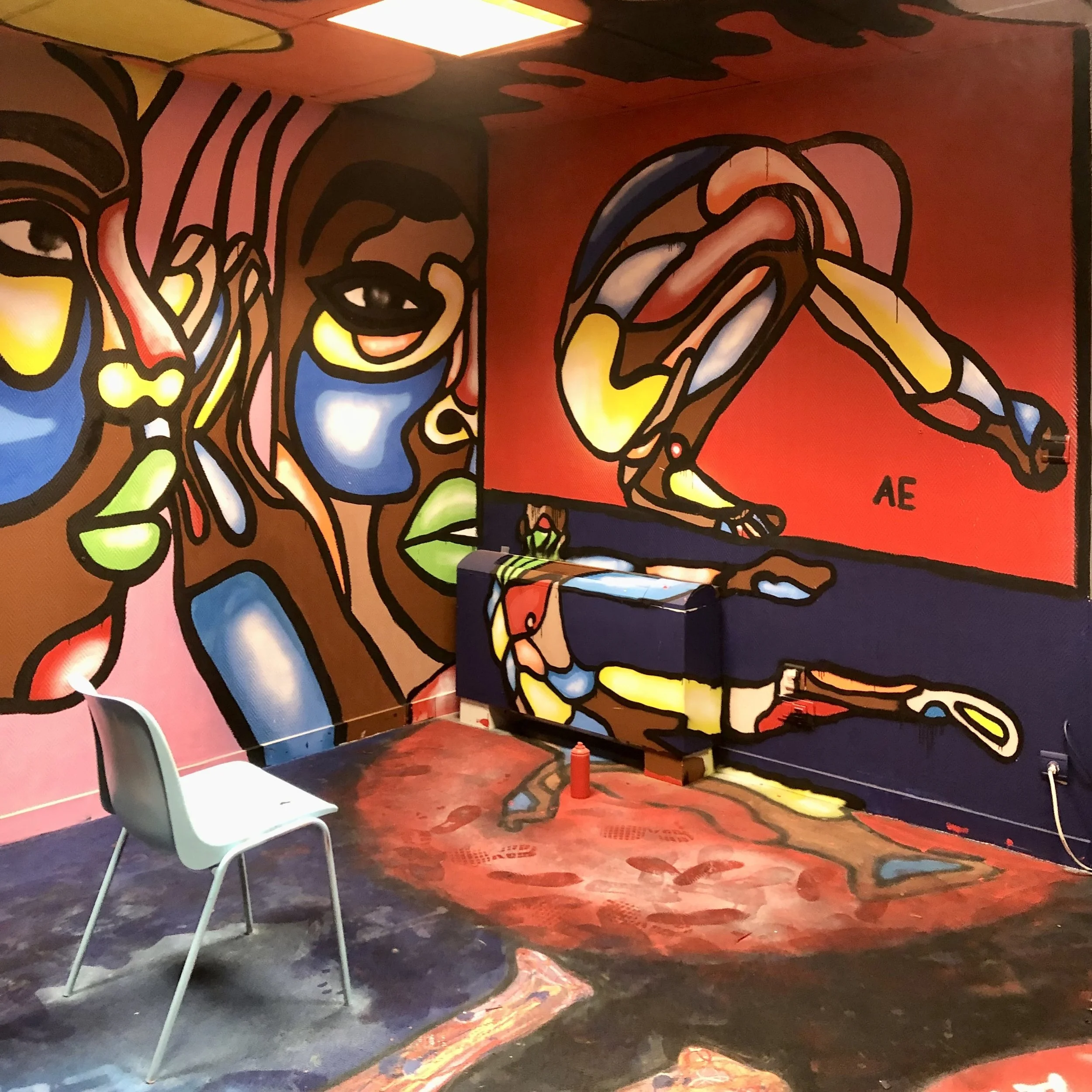 Un mur peint avec une œuvre street art abstraite colorée représentant des visages stylisés et une figure humaine en position de squat, dans un décor coloré et artistique.