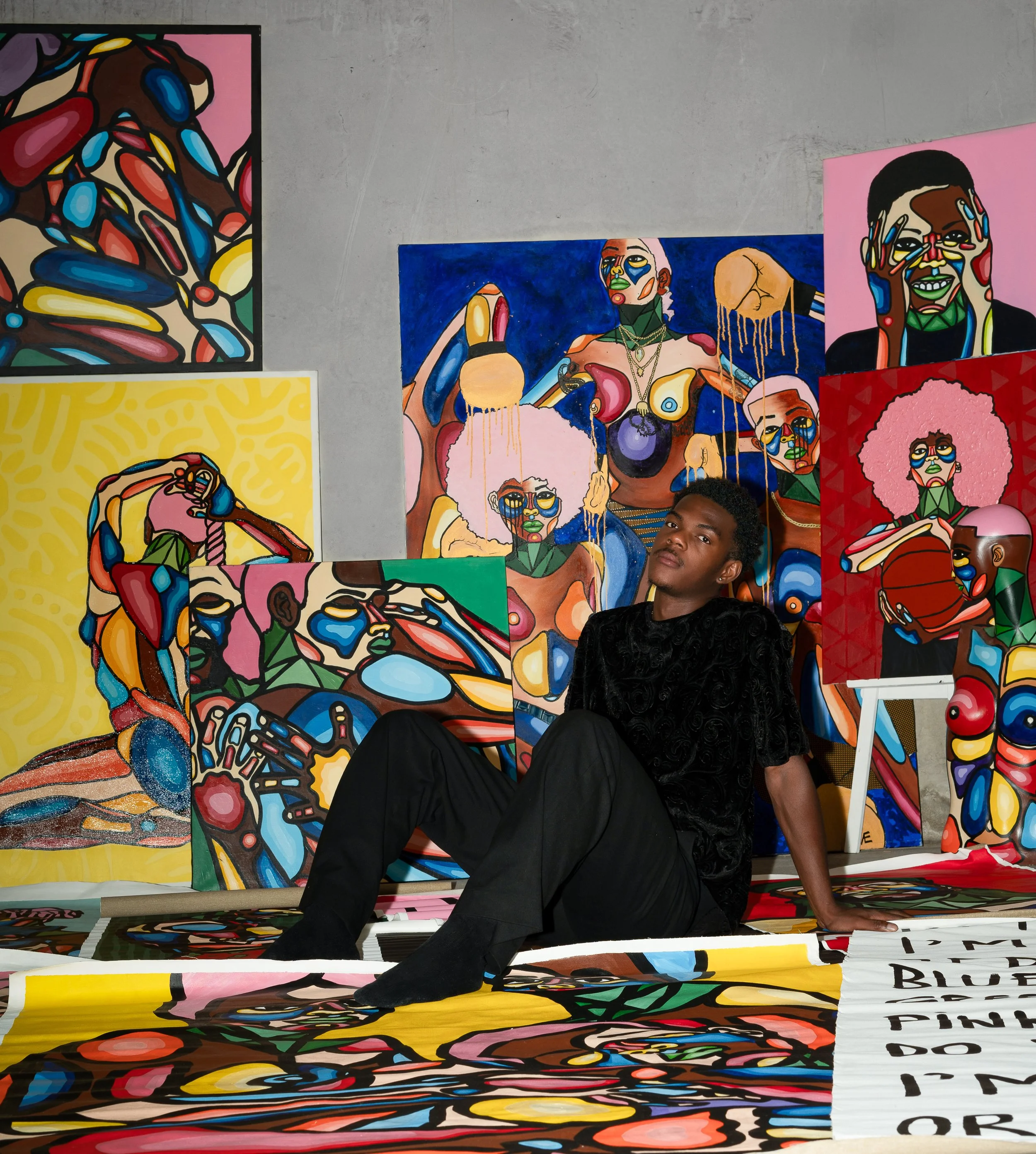 Un jeune homme assis devant plusieurs œuvres d'art colorées et abstraites représentant des personnages avec des visages expressifs.