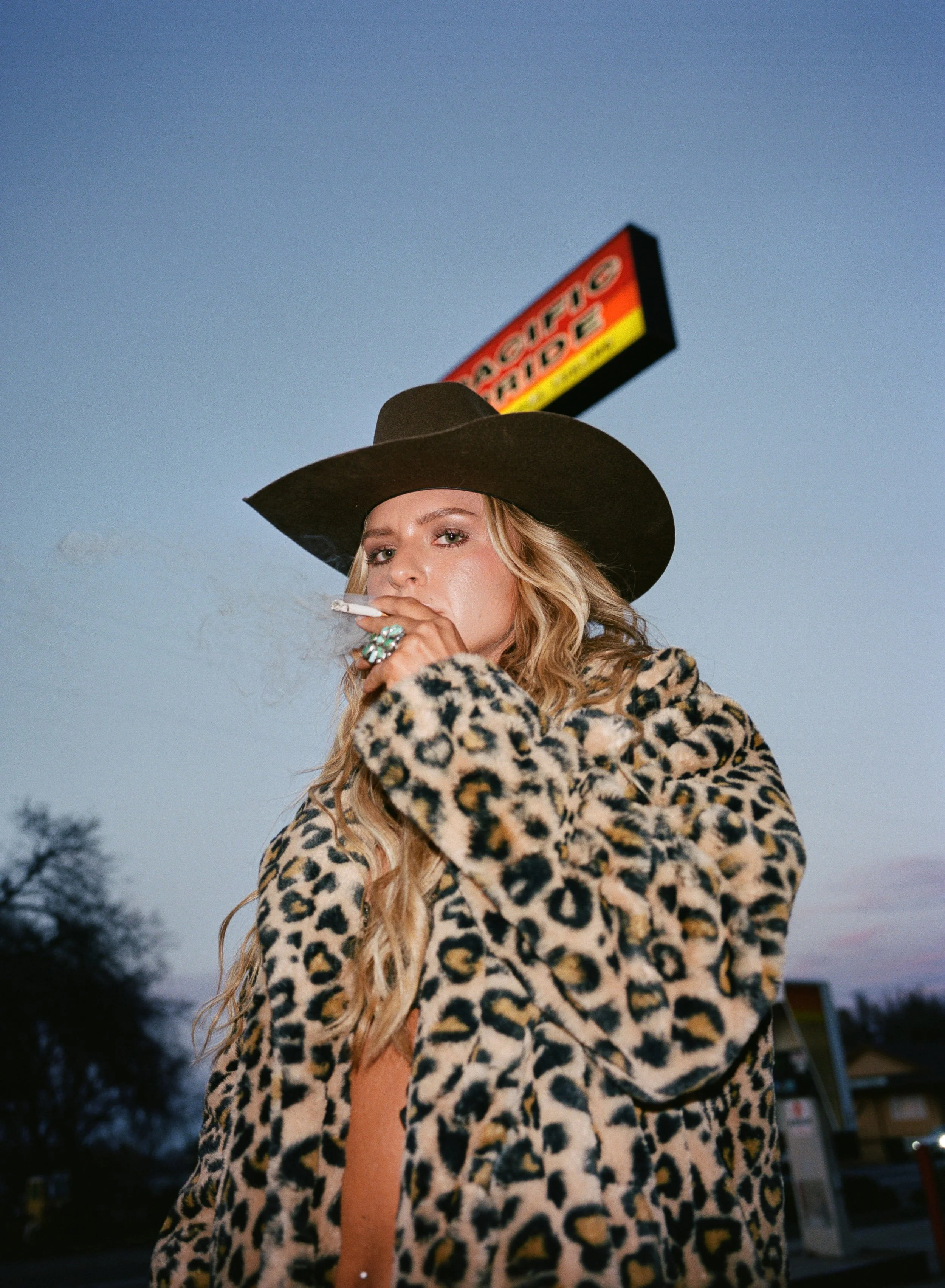 A woman wearing a leopard print coat and a large black hat, smoking a cigarette outdoors during dusk, with a sign in the background and trees in the distance.