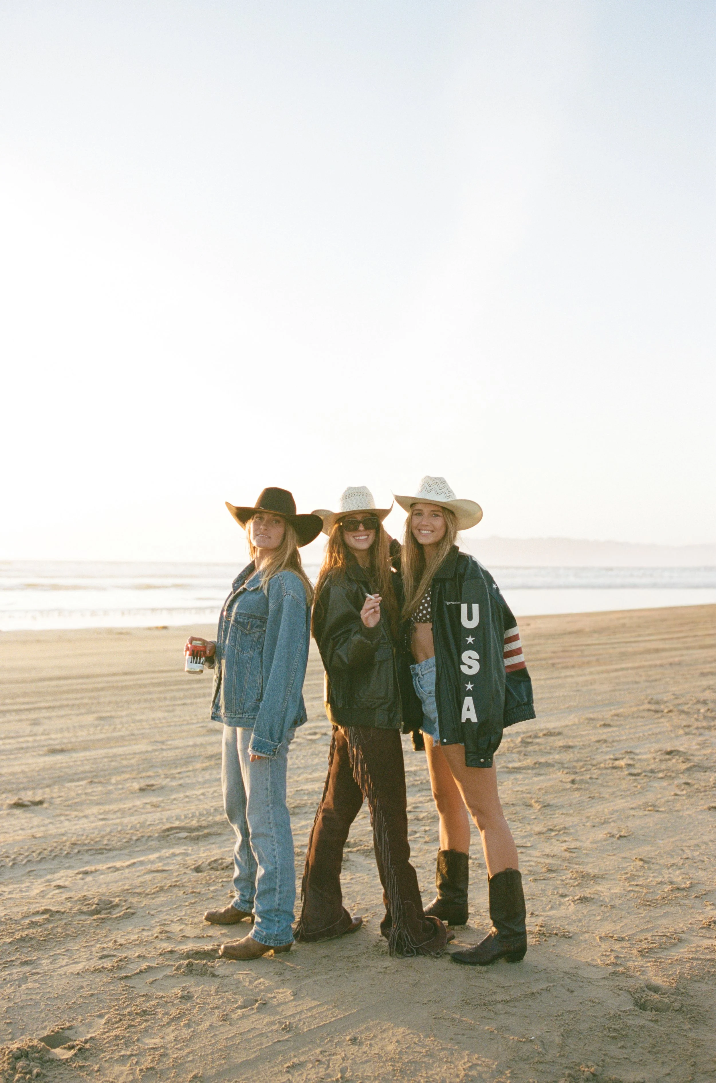 Three young women standing on a beach at sunset, wearing cowboy hats and casual clothing, smiling and enjoying the outdoors.