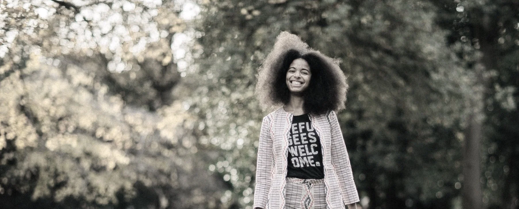 A smiling person with an afro standing outdoors among trees, wearing a patterned blazer and pants with a black T-shirt that has white text.