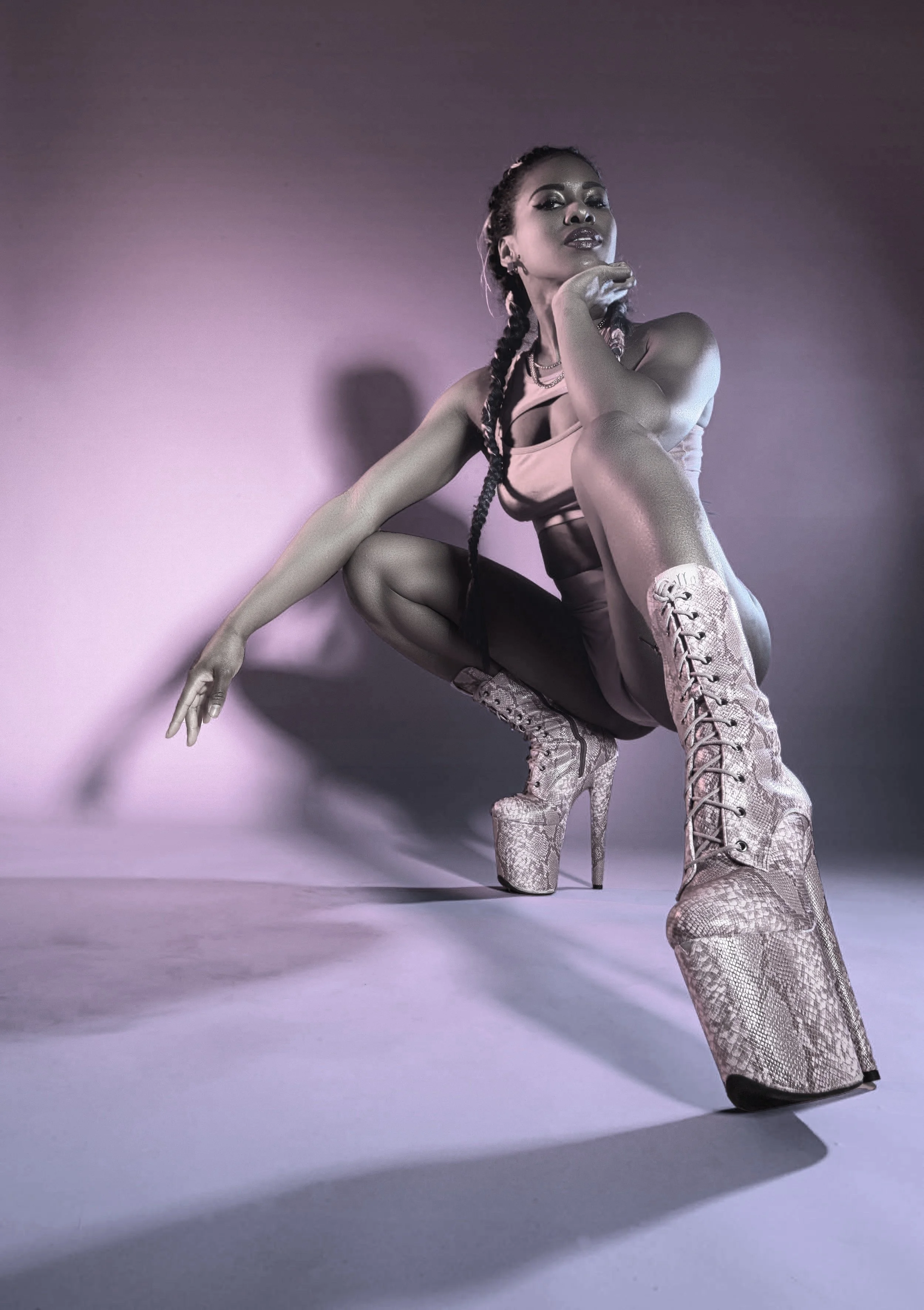 A person with braided hair poses against a pink and purple gradient background, wearing high-heeled lace-up boots and a beige bodysuit.