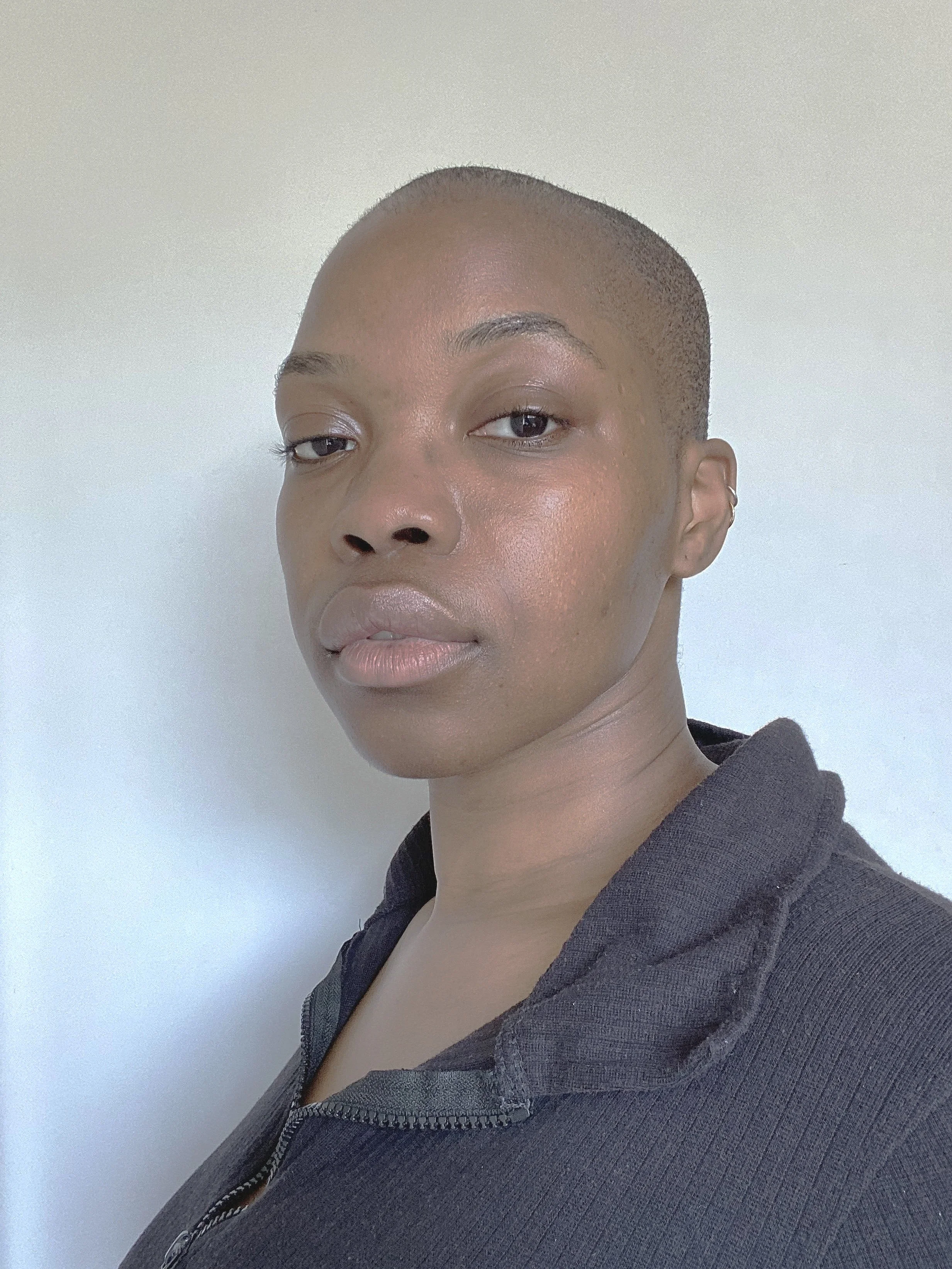 Close-up of a person with a shaved head, light skin, and wearing a dark grey zip-up hoodie against a plain light-colored background.