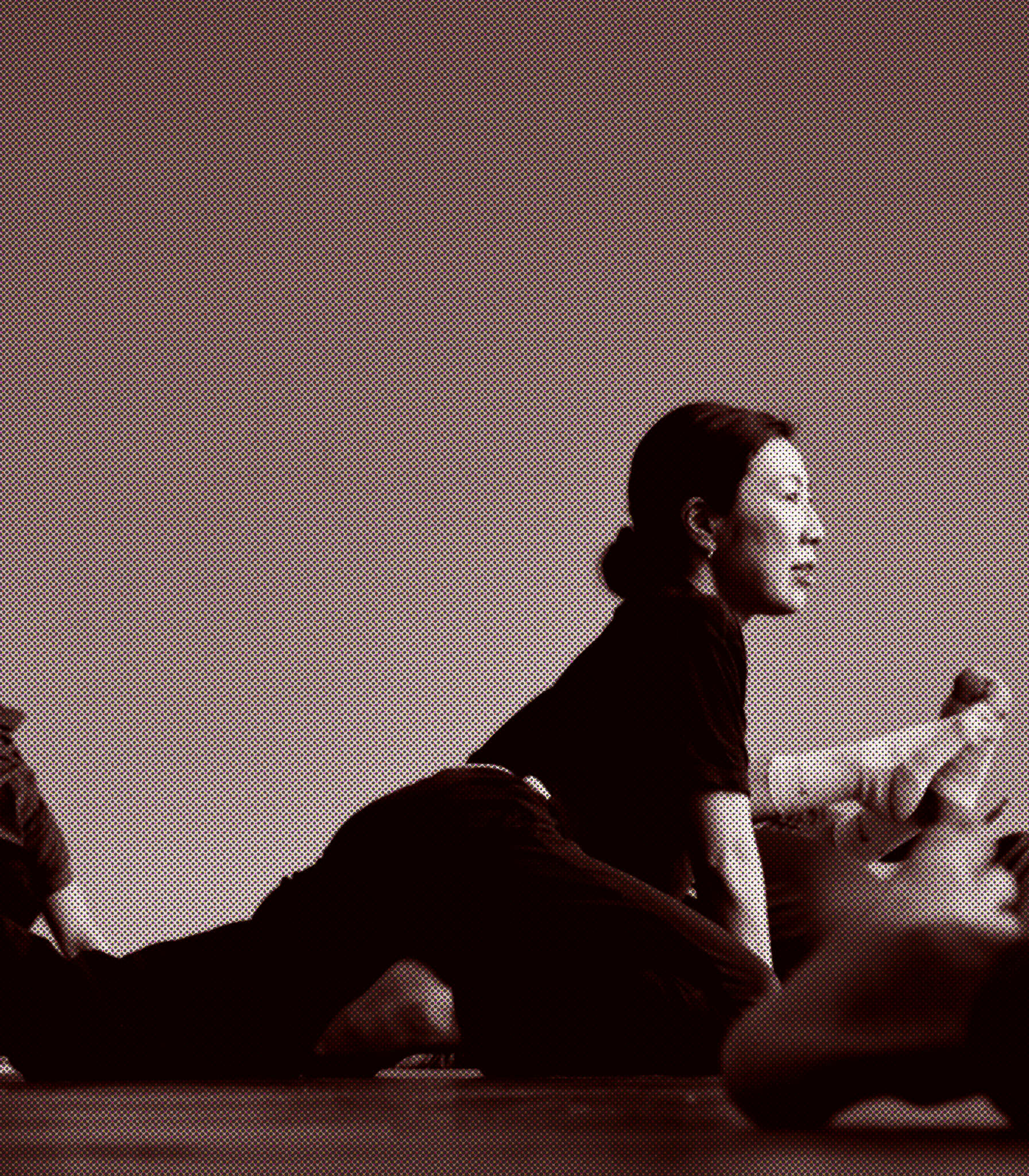 A woman with dark hair tied back, sitting at a table with her eyes closed and hands raised, possibly meditating or practicing mindfulness, in a softly lit indoor setting.