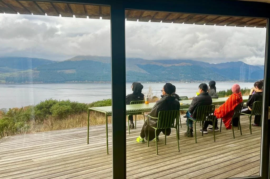People sitting on a wooden deck and looking out at a lake and mountains in the distance.