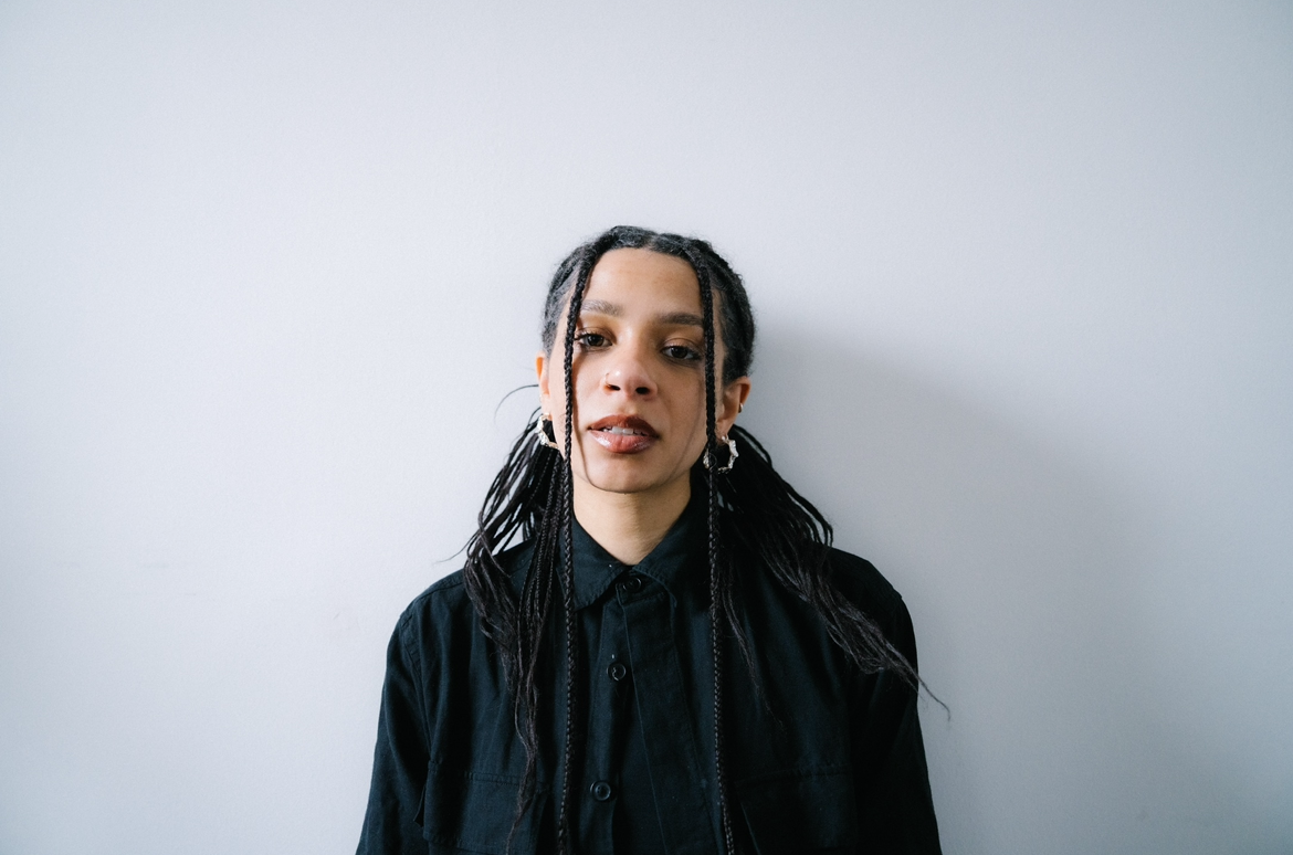 A woman with braided hair wearing earrings and a black button-up shirt, standing against a plain white wall.