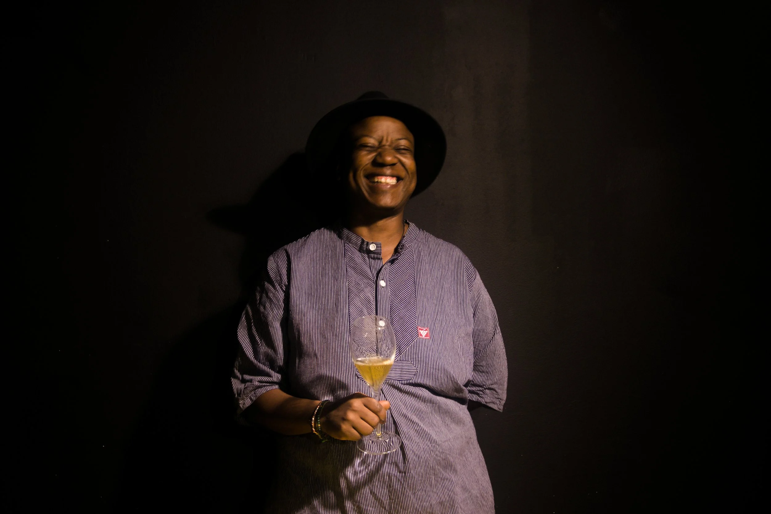 A man smiling and holding a glass of champagne, wearing a striped shirt, a black hat, and various bracelets, standing against a dark background.