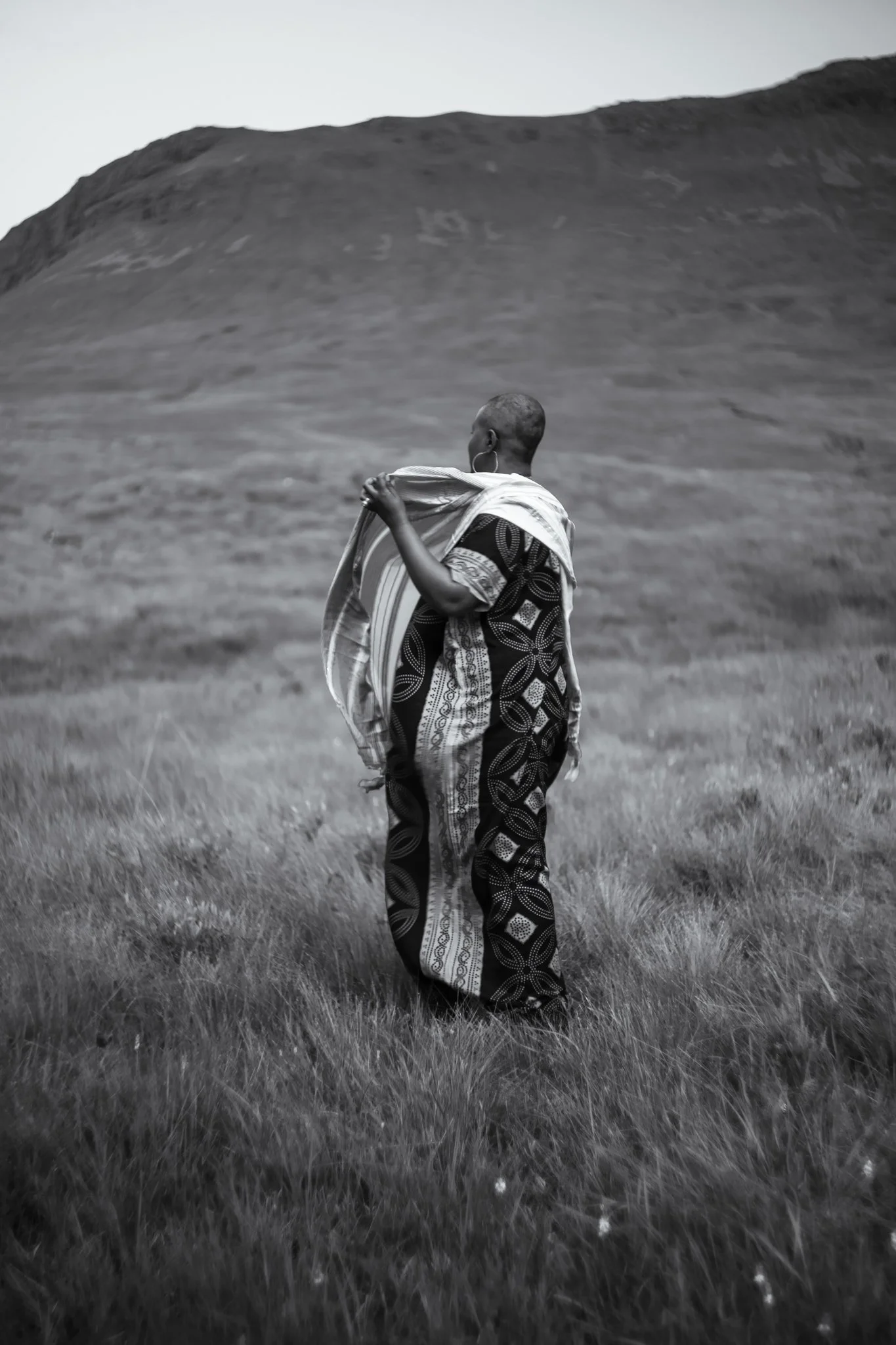 A black and white photo of a person walking through a grassy landscape with large hills or mountains in the background. The person is wearing traditional clothing and earrings, holding a piece of cloth over their shoulder.