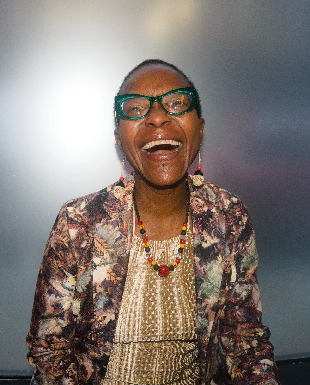 A joyful woman with short hair and glasses, wearing colorful earrings and a patterned jacket with a beige top, smiling widely.