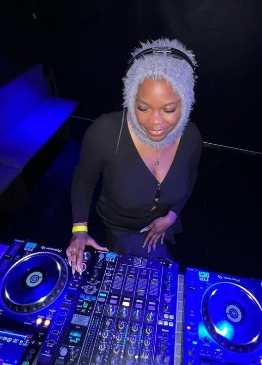 A woman with curly white hair and headphones is DJing at a club, standing behind a professional DJ setup with turntables and a mixer, illuminated by blue and purple lighting.