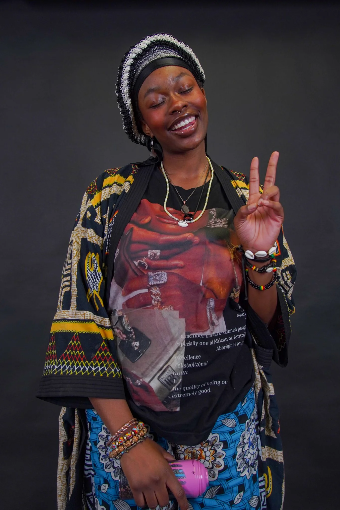 A woman with dark skin and multiple colorful beaded bracelets on her arm smiling with her eyes closed and making a peace sign with her fingers. She is wearing a patterned jacket, a graphic t-shirt, a beaded headscarf, and layered necklaces, standing against a plain dark background.
