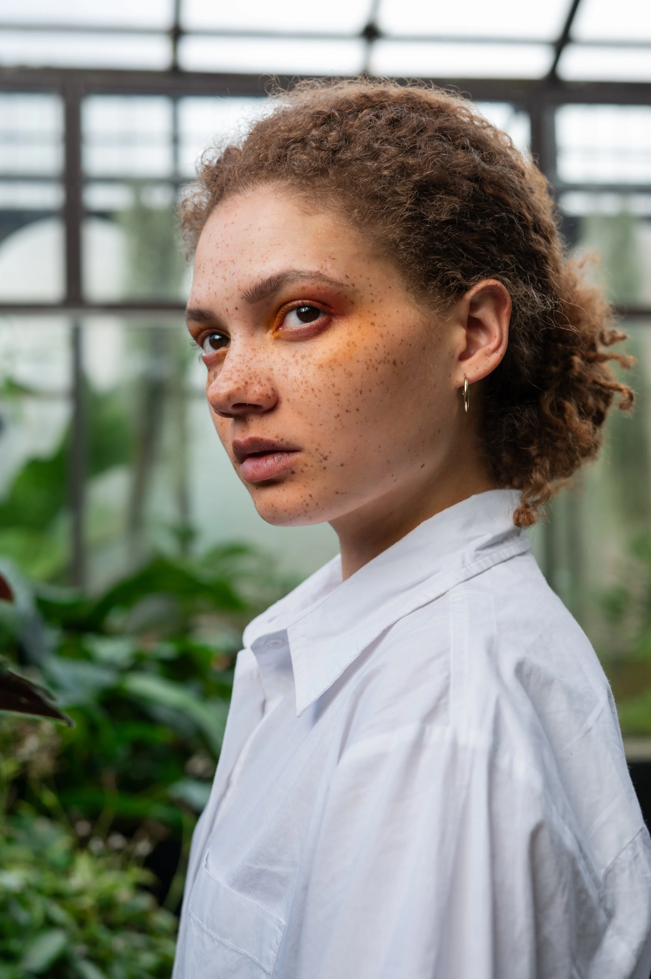 Close-up of a young woman with curly hair and freckles, wearing a white shirt and a hoop earring, standing in a greenhouse with green plants and glass roof in the background.