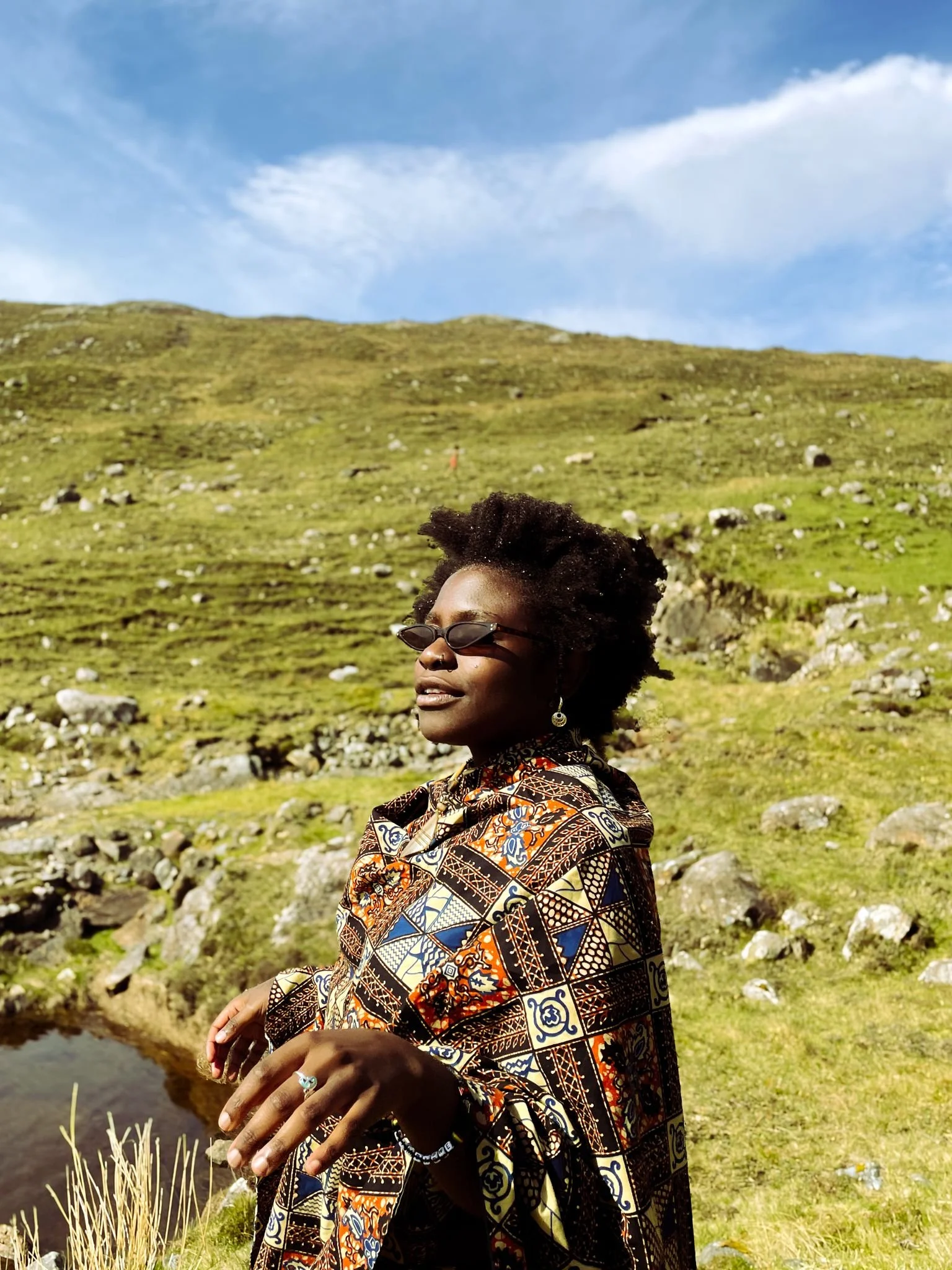 A woman with dark skin, wearing sunglasses and large earrings, dressed in a patterned African print dress, standing outdoors in a green hilly landscape with rocks and a pond, under a blue sky with scattered clouds.