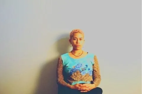 A woman with short blonde hair sitting against a plain light-colored wall.