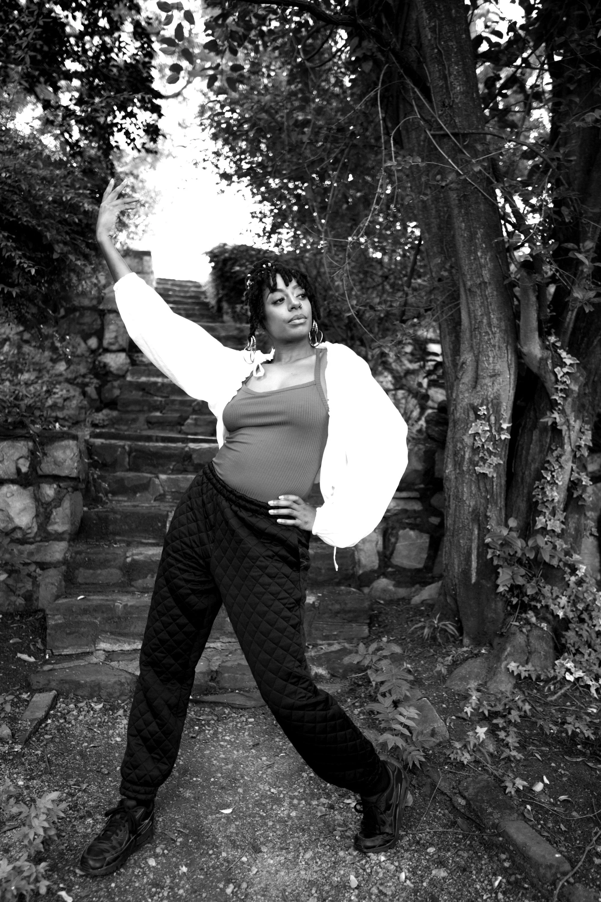 A woman with dreadlocks poses outdoors on stone steps surrounded by trees and bushes, wearing a tank top, quilted pants, and a jacket.