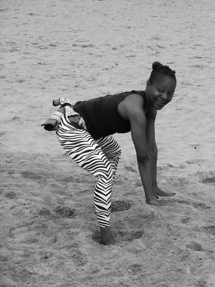 A woman in striped pants standing on a sandy beach, smiling, with her hands and one foot on the ground and the other leg bent back with the foot held by her hand.