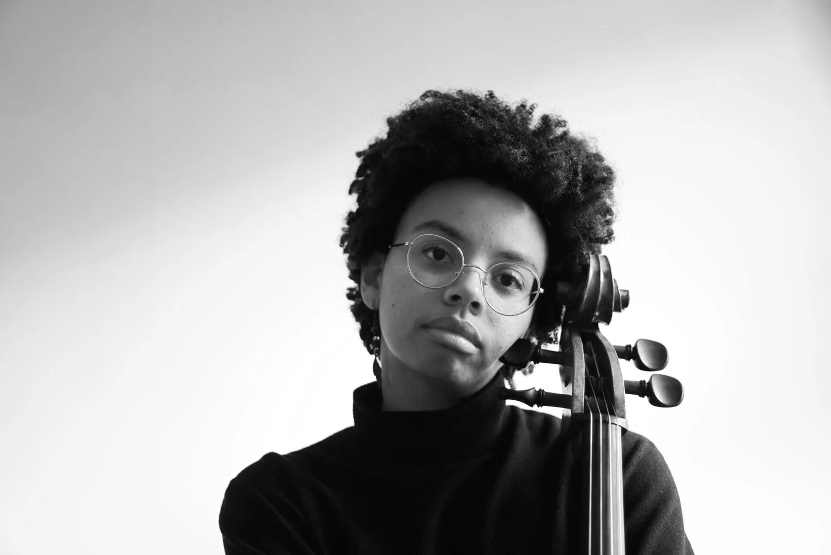 Black and white portrait of a young woman with an afro, wearing glasses, holding a cello, and looking directly at the camera.