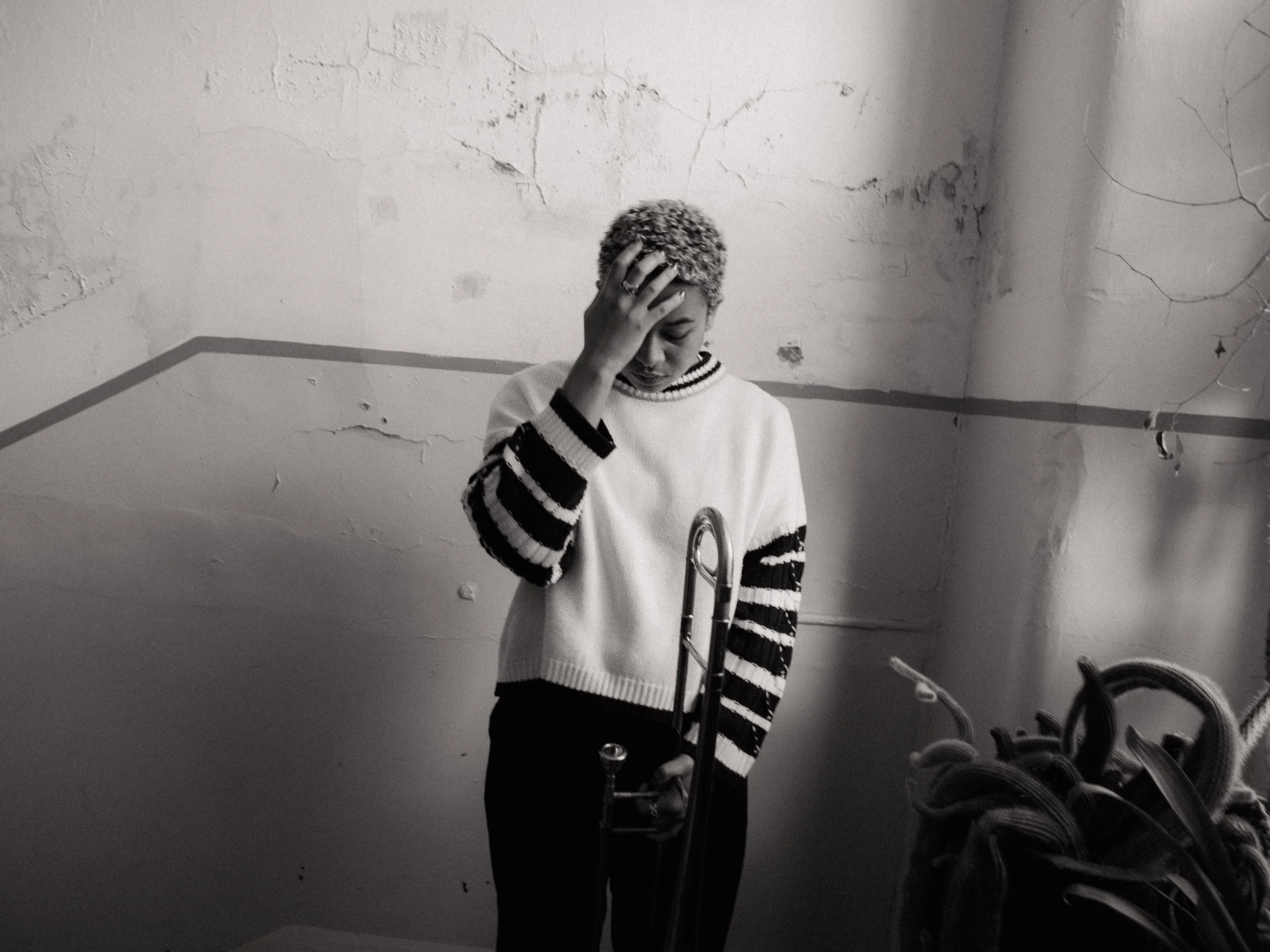 A person with short, curly hair and wearing a striped sweater is standing in front of a mirror, holding their head with one hand. The background shows a cracked, textured wall and a line painted across it. There are bicycles partially visible in the lower part of the image.