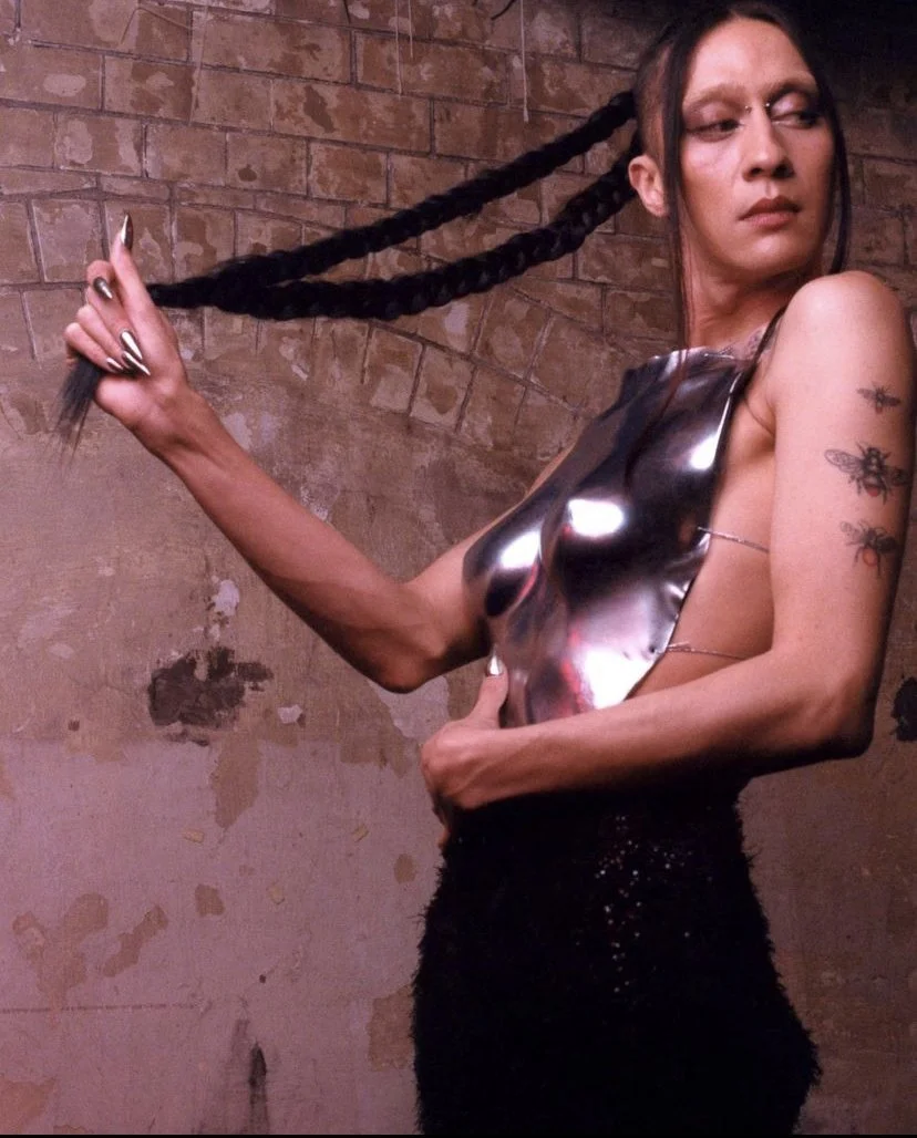 A woman with tattoos on her right arm, wearing a reflective metallic top and black skirt, is holding a long braid of her black hair against a brick wall background.