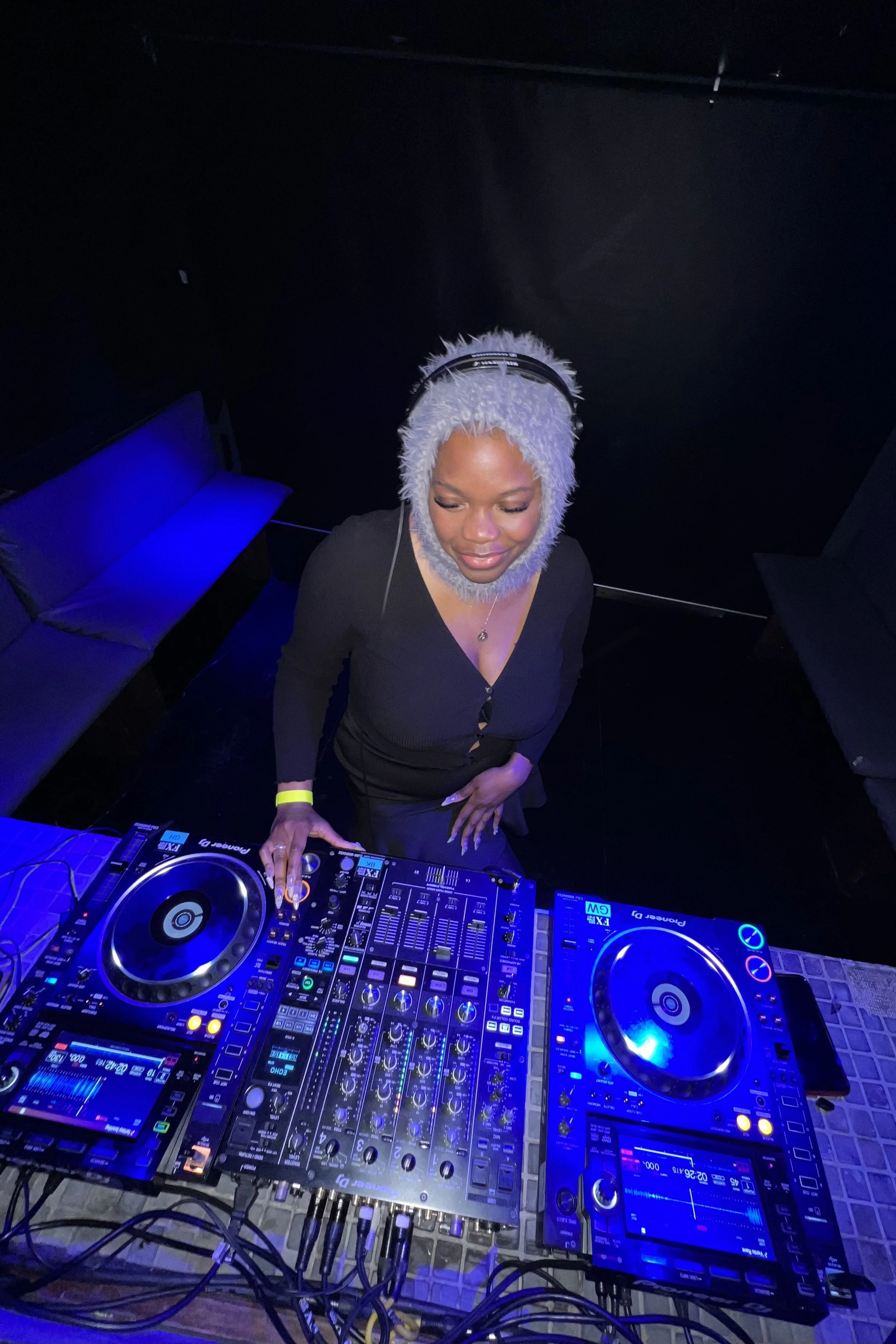 A woman with a fluffy gray hood over her head, wearing a black top, is DJing at a nightclub with a professional DJ controller in front of her. The club has dark decor with blue lighting.