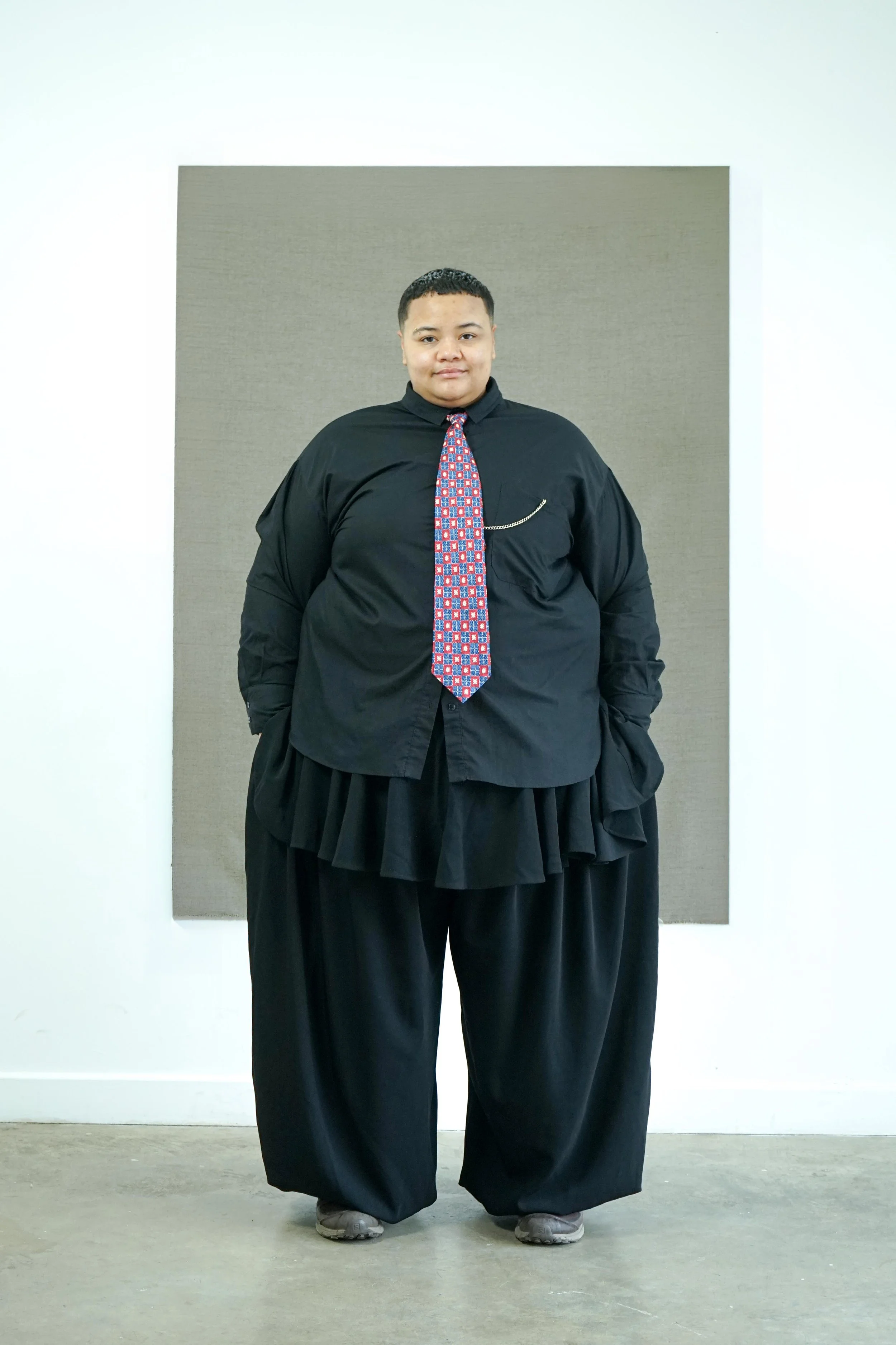 A person standing in front of a neutral background, wearing a black shirt, a colorful patterned tie, and oversized black pleated trousers. They are also wearing gray sneakers.