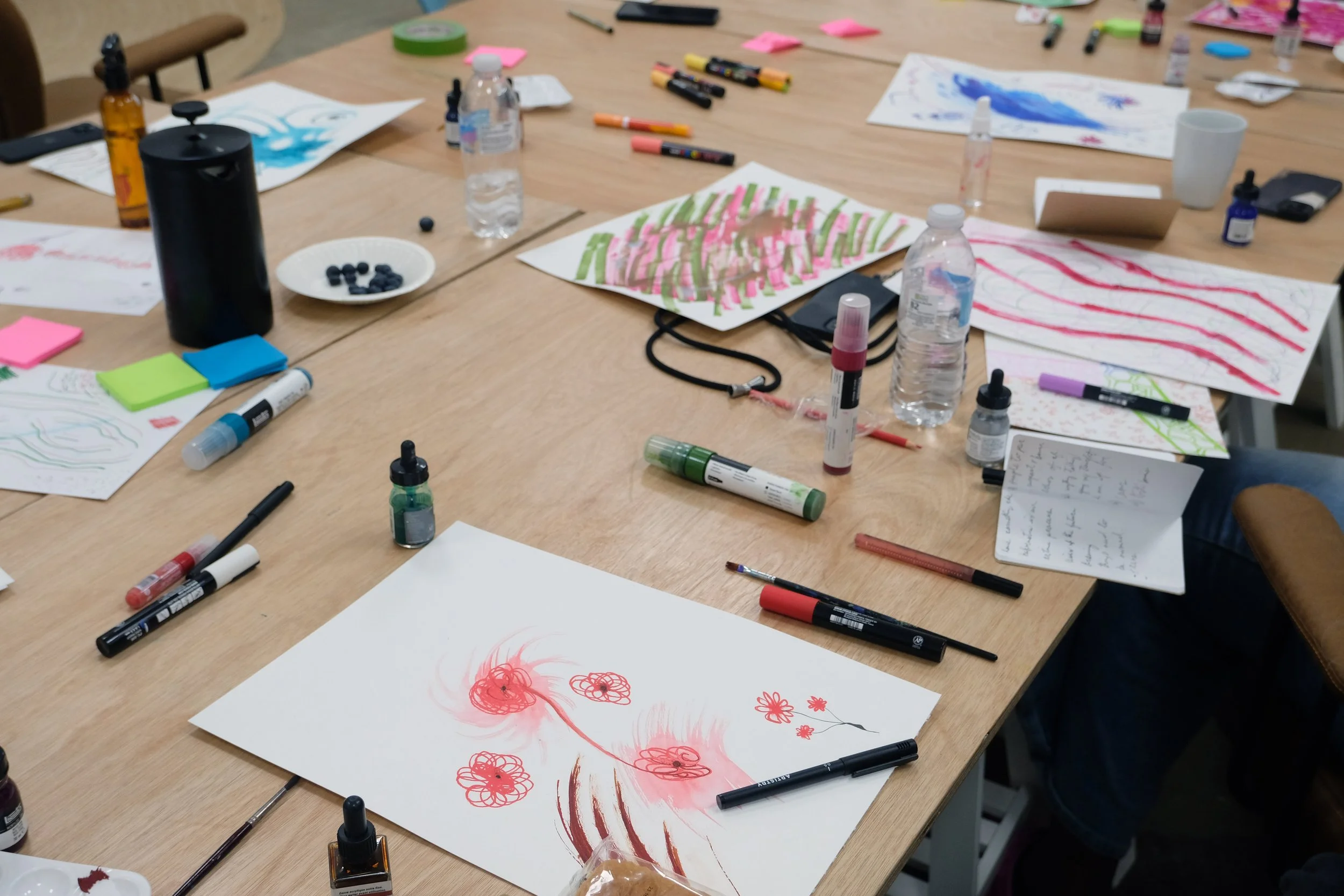 A cluttered table with art supplies including markers, watercolor paints, papers with drawings of flowers, a water bottle, sticky notes, and other art tools.