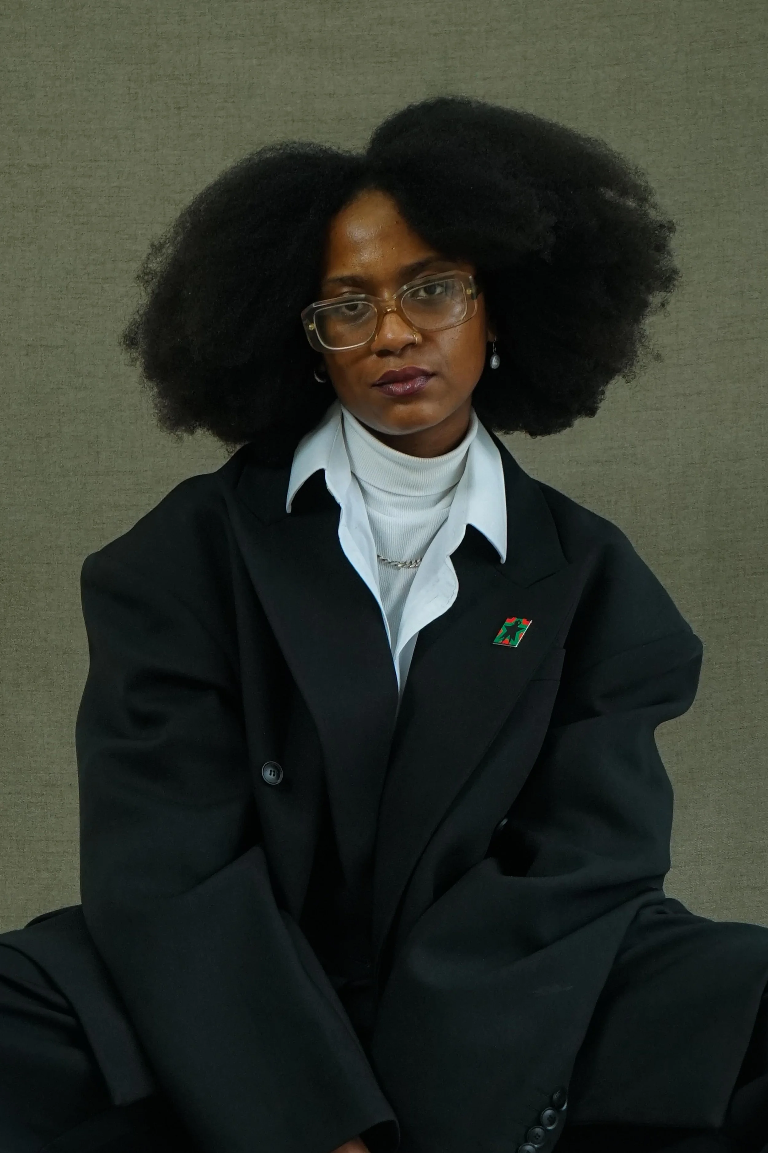 A person with a black afro, glasses, and earrings, wearing a black blazer and white shirt, sitting against a plain beige wall.