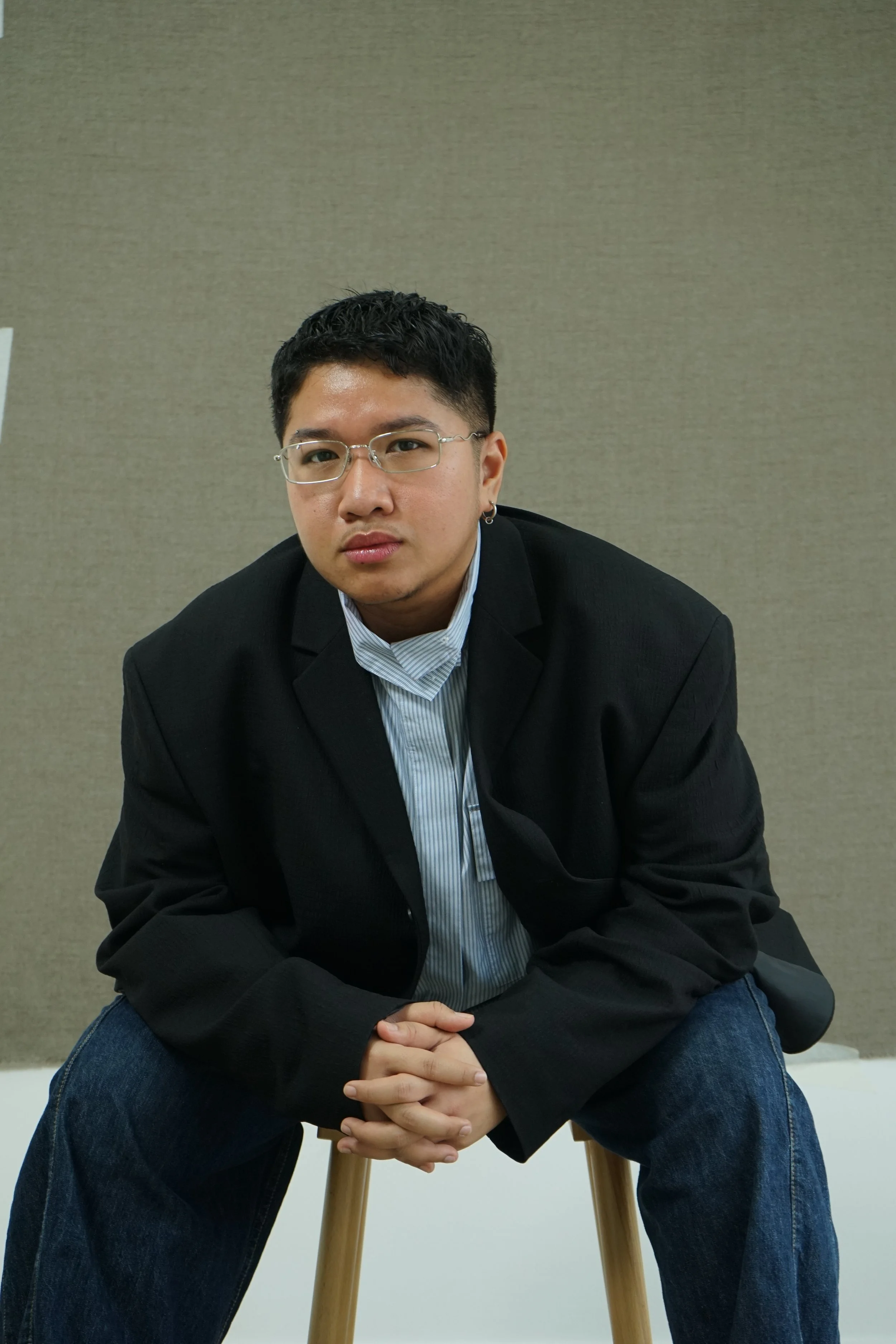A person with glasses and an earring, wearing a black jacket and striped shirt, sitting on a wooden stool with a beige background.