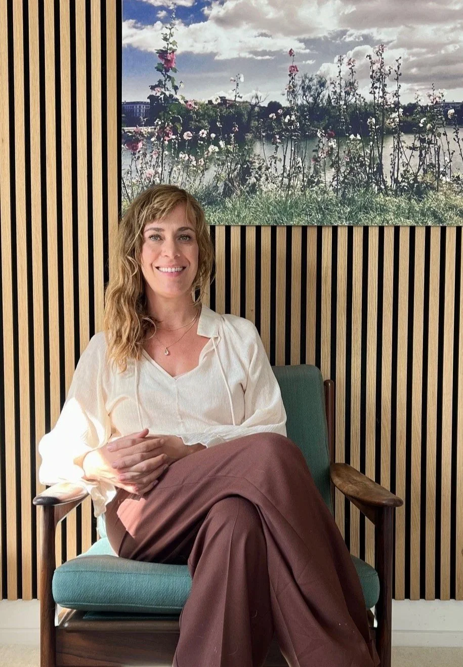 A woman with wavy blonde hair, wearing a cream blouse and brown pants, sitting on a green armchair in front of a wooden slat wall with a large window showing a river, trees, and flowers.