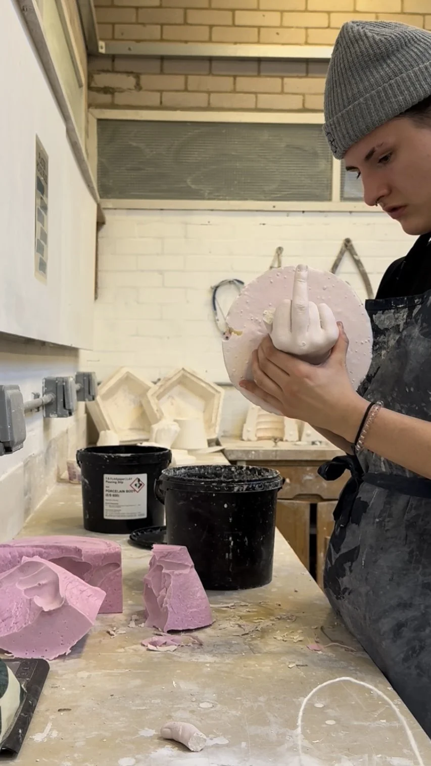 A person working in a pottery studio holding a pink ceramic piece and showing a middle finger gesture.