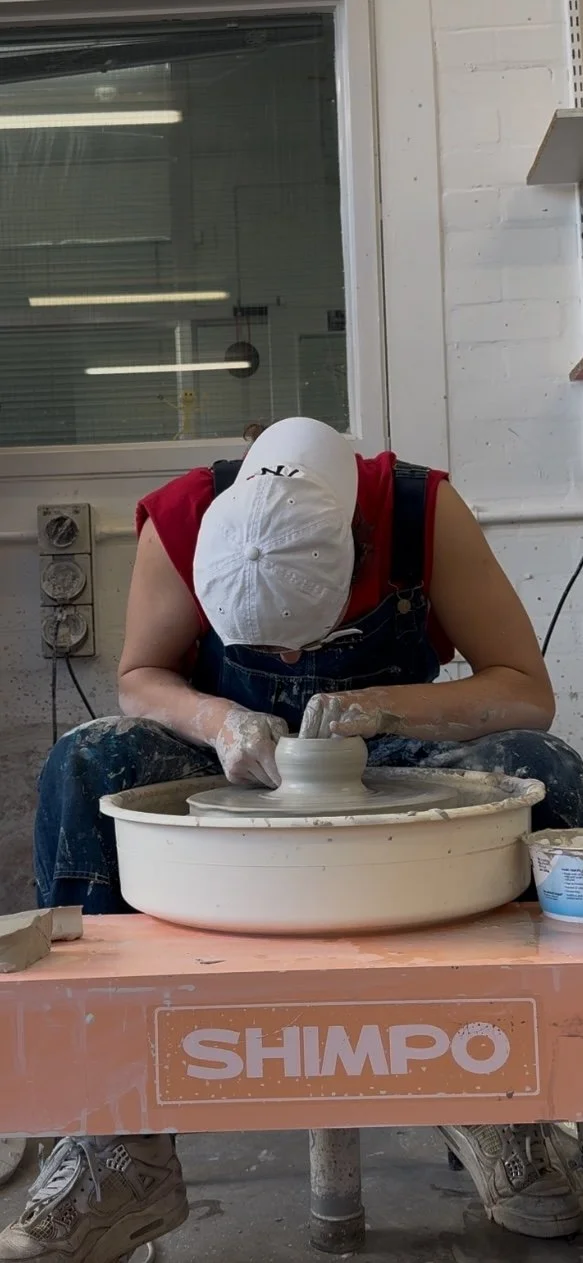 Person wearing a white cap and red sleeveless shirt working on a pottery wheel in a workshop.