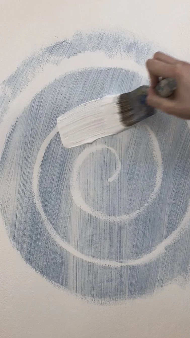 A person hand holding a wide paintbrush painting white concentric circles on a wall, creating a target-like design.