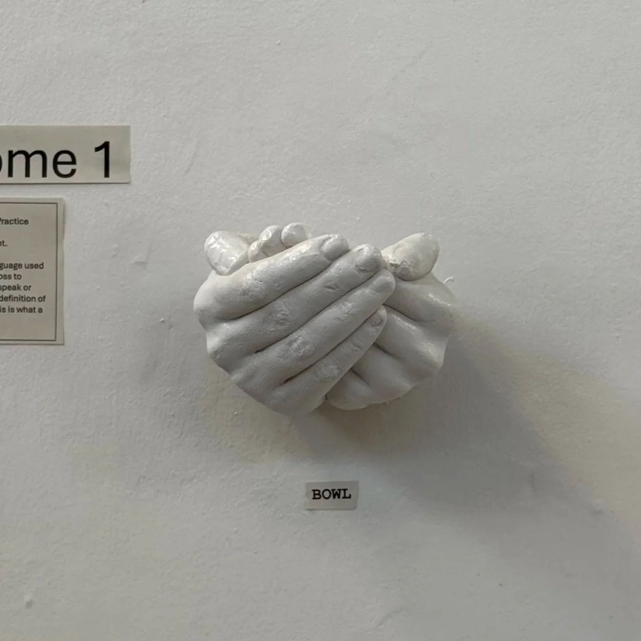 A white sculpted art piece showing two hands 'signing' bowl, mounted on a white wall with a small label underneath that says "BOWL."
