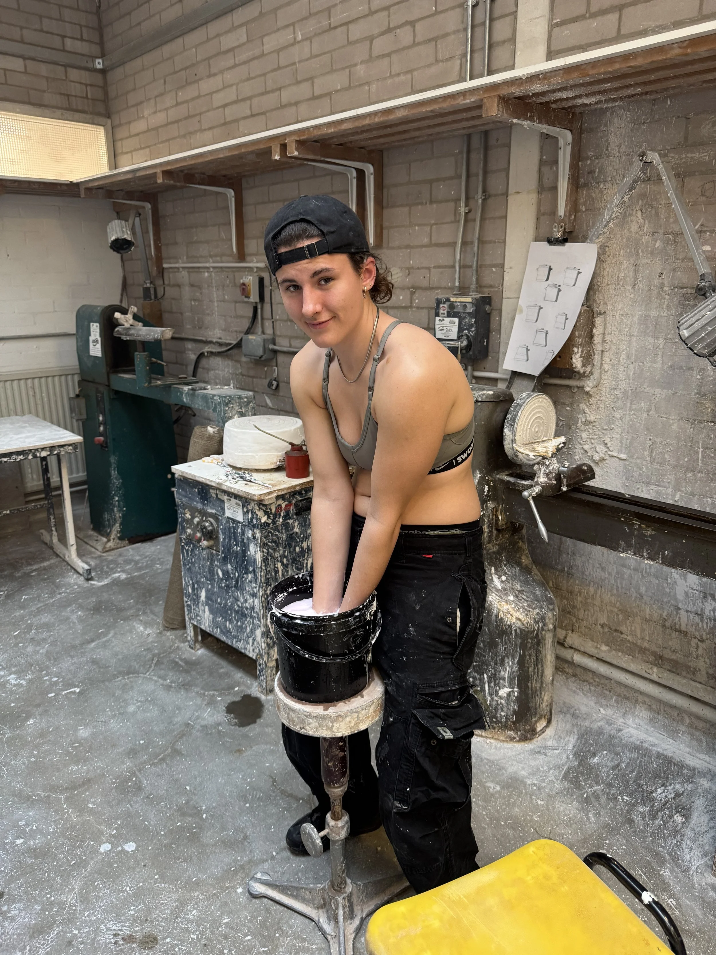 A young woman in a gray sports bra and black cargo pants stands in a pottery workshop, mixing or kneading clay with both hands inside a black bucket, with a slight smile. She wears a black backward cap and has a silver necklace, surrounded by pottery