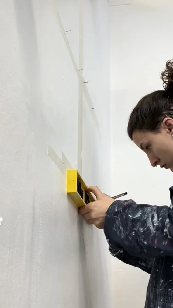A person with dark curly hair, wearing a dark paint-stained shirt, using a level on a wall.