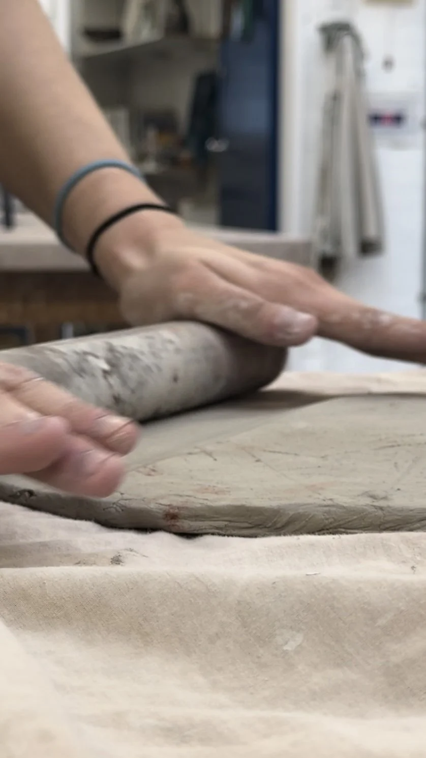 A person rolling out clay with a cylindrical rolling pin in a kitchen.