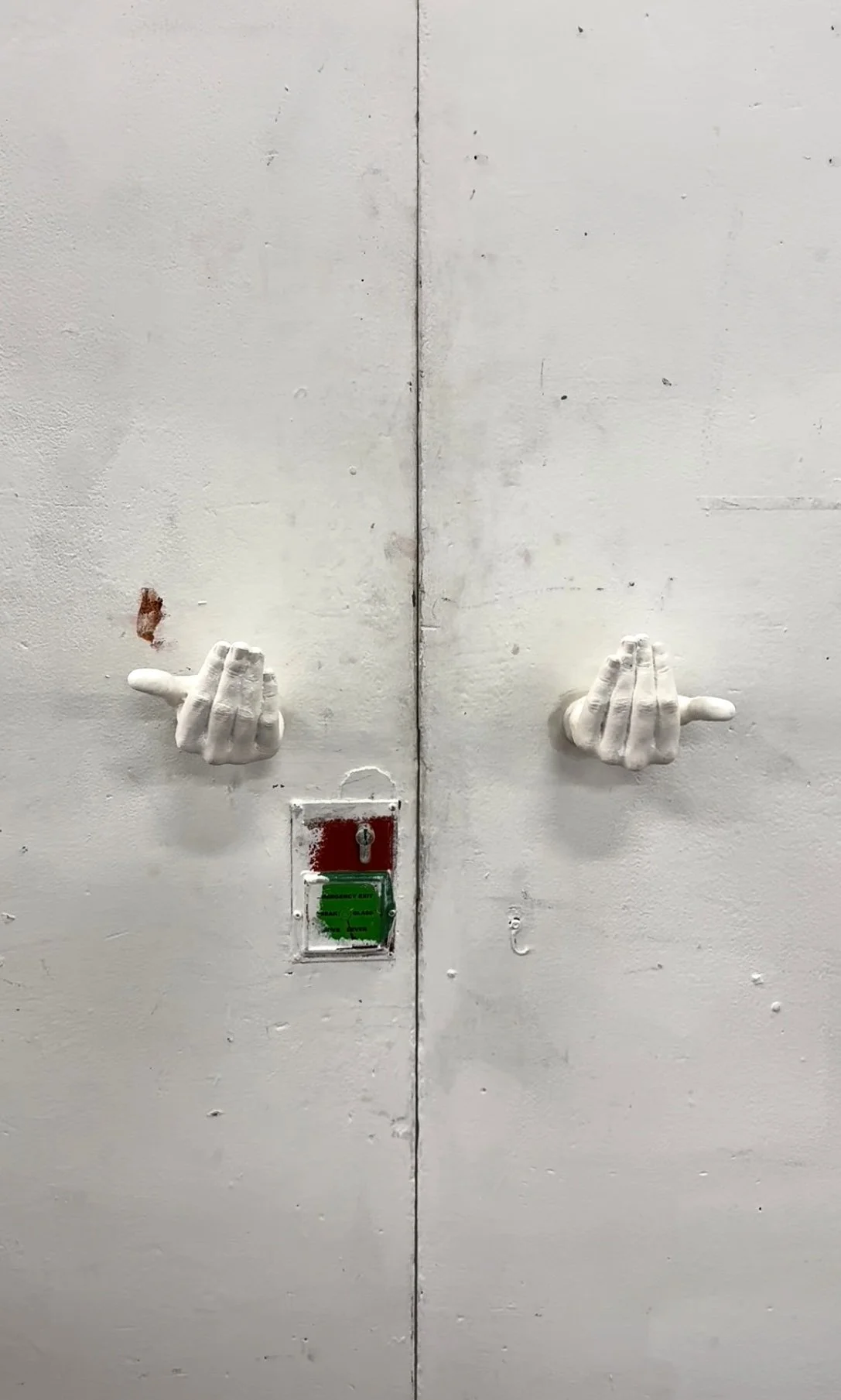 Two white mannequin hand sculptures extended from a white wall, positioned on either side of a small, locked access panel with a red and green indicator, on a worn, painted surface.