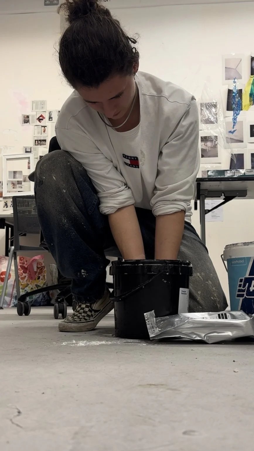 Person crouching on the floor with paint on clothes, reaching into a black bucket surrounded by painting supplies, in an art studio.