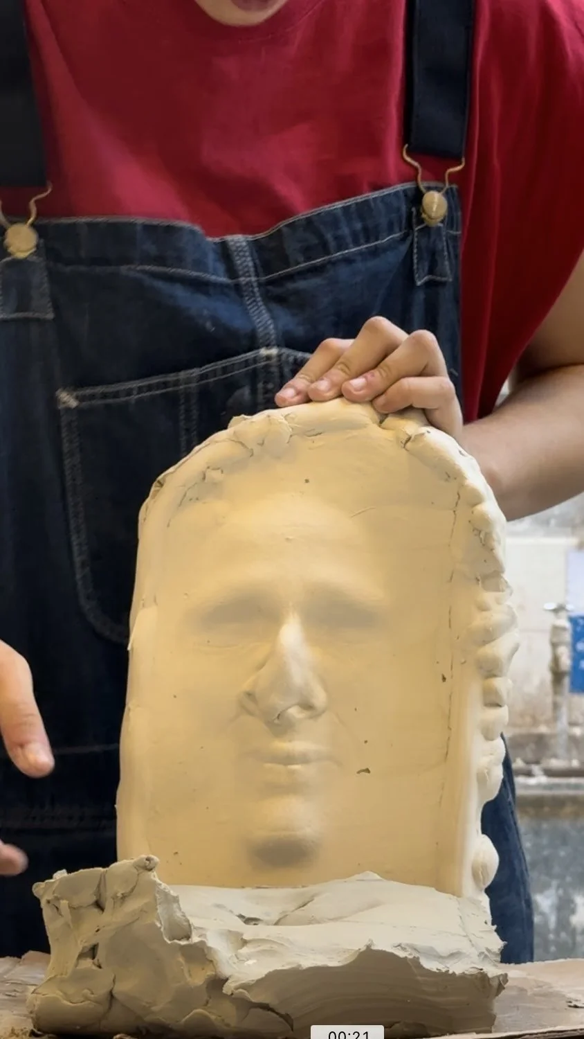 A person in a red shirt and denim overalls holds a large sculpted face made of clay.