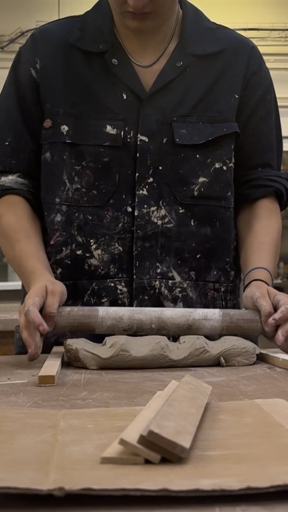 Person in a black paint-splattered shirt rolling out clay with a rolling pin in a woodworking or pottery studio.