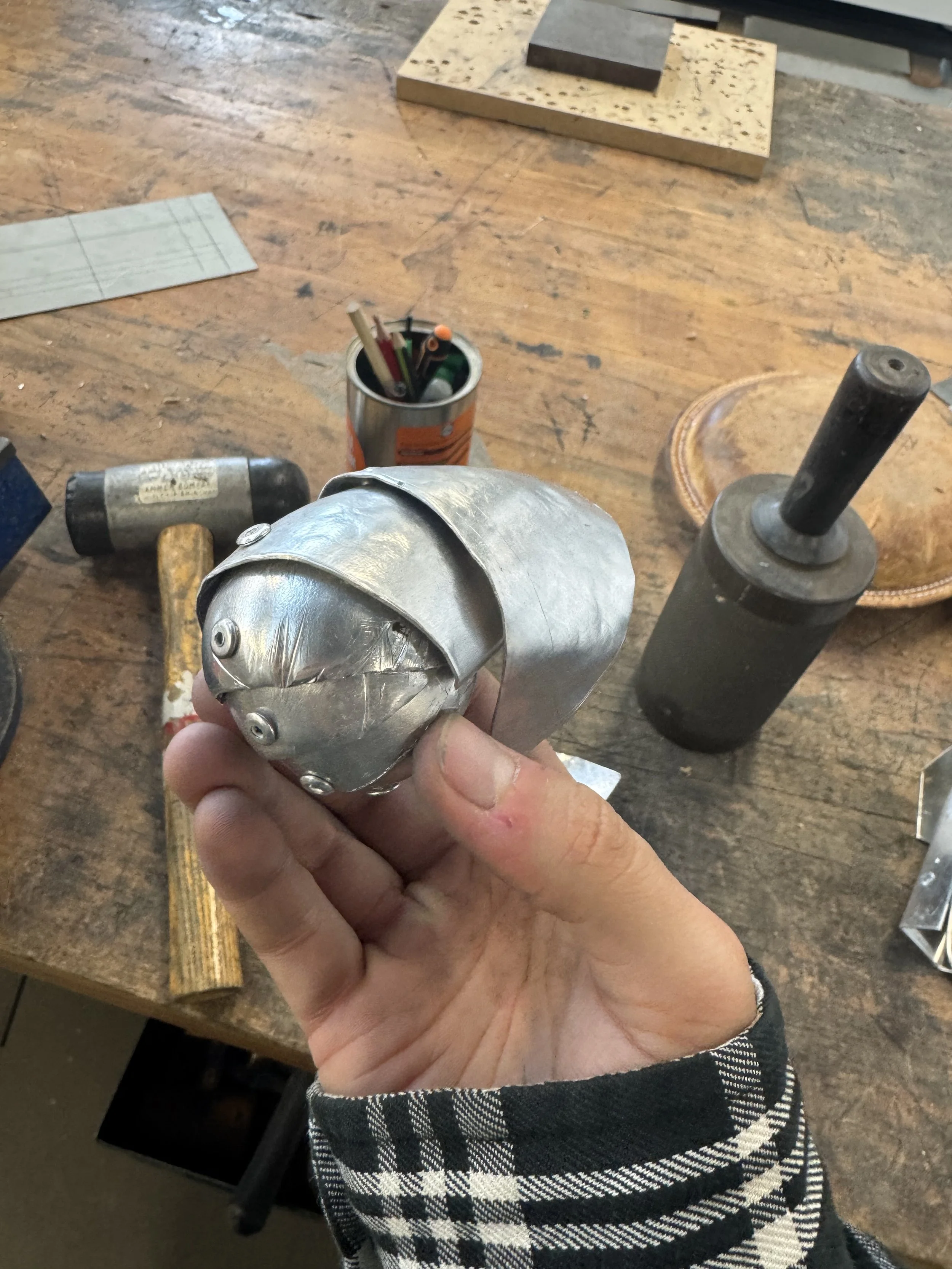 A person holding a small, shiny metallic object resembling a robot or a futuristic device, in a workshop setting with tools, a hammer, and samples of materials on a wooden workbench.