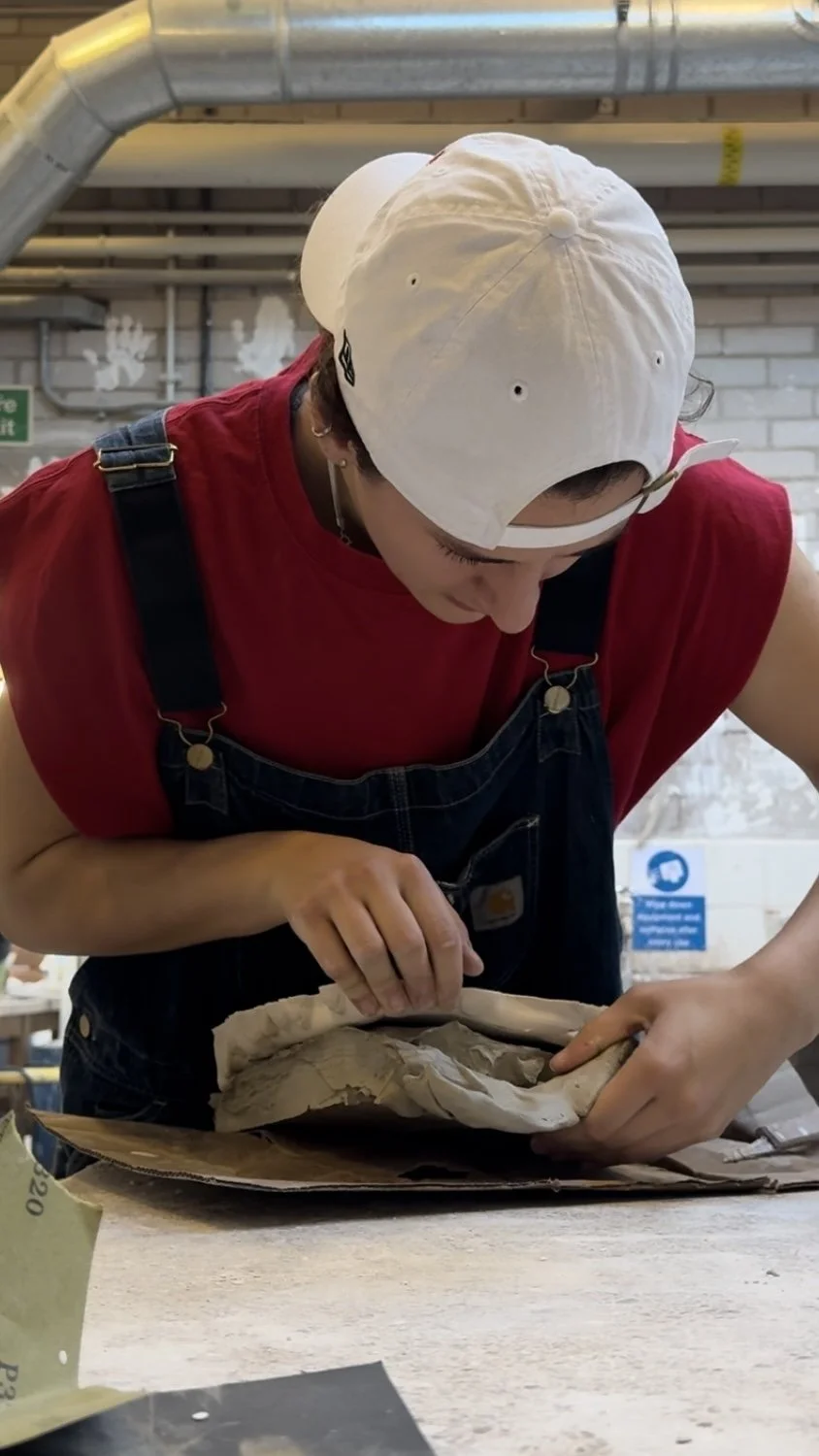 A person wearing a white cap and red sleeveless shirt with overalls, working with clay or ceramic on a table in a workshop.